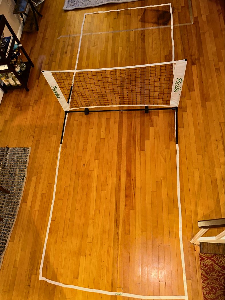 A mini indoor volleyball net set up on a hardwood floor, with white tape marking out a court boundary. The net has white side panels with green 'Pasta' branding.
